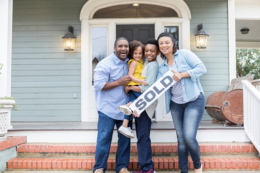 Attractive family smile proudly while holding a 'SOLD' sign in front of their new home.