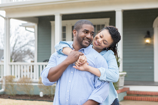 Loving mid adult African American man and his beautiful wife embrace while standing in front of their new home.