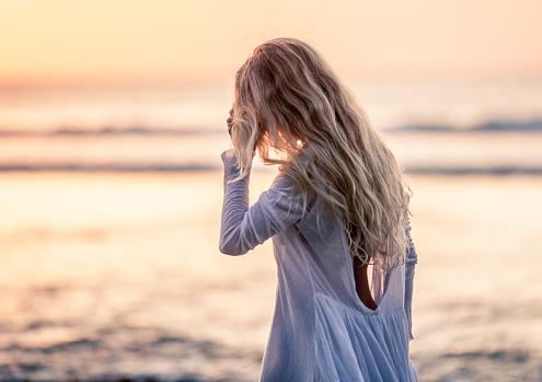 Beautiful blonde girl with long hair in short white dress dancing at sunset on the beach in Bali, Indonesia