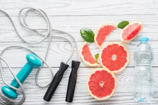 Jumping-rope, dumbbell,  fresh grapefruits and bottle of water, on rustic white wooden table , top view, flat lay, fitness accessories. Concept of slimming, dieting and healthy nutrition.