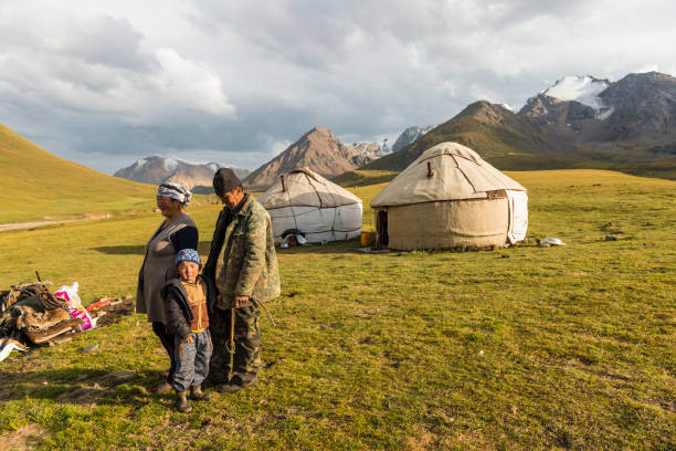 Scenic view on nomad family, women, man and a boy at lake Kol-Ukok against two yurts and green hill and mountain range on sunny day.