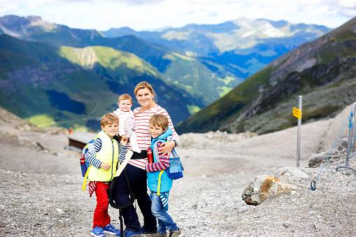 Happy mother with little baby girl and two kids boys travelling in backpack. Hiking adventure with three children in mountains. Vacations journey weekend travel in Hintertux glacier, Austria
