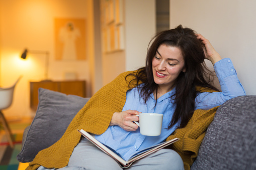 Brunette woman sitting on sofa , drinking tea and reading book at her home