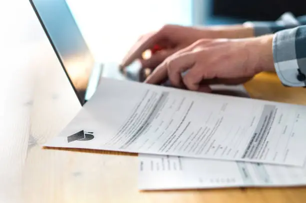 Photo of Young man writing college or university application form with laptop. Student applying to school. Scholarship document, admission paper or letter on table. Typing email.