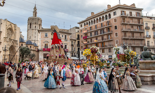 March 17, 2018. Valencia, Spain. In fallas festivity, there's an event that stands out above all others, it is the floral offering to the Virgin. All the falla groups meet at their headquarters, dressed in traditional costume, and they walk on the streets, accompanied by musical groups and carrying bunches of flowers to be placed at the feet of the figure of the Virgin. It is a stunning ceremony that converts the central city square into a wonderful flower show. Participants carry bouquets of flowers with lots of pageantry and applause, because they simply love seeing and being seen, and ladies' costumes are very much a part of the picture. Virgin square is transformed into a flower garden during the offering. The façade of the basilica becomes a floral tapestry that covers the entire wall, giving shape to a huge representation of Our Lady, and in the centre of the square, a 14m high wooden structure is set up, on which agile climbers insert the flower offerings as they are thrown up to them, creating a gigantic effigy of Virgin Mary. People look and enjoy the event.