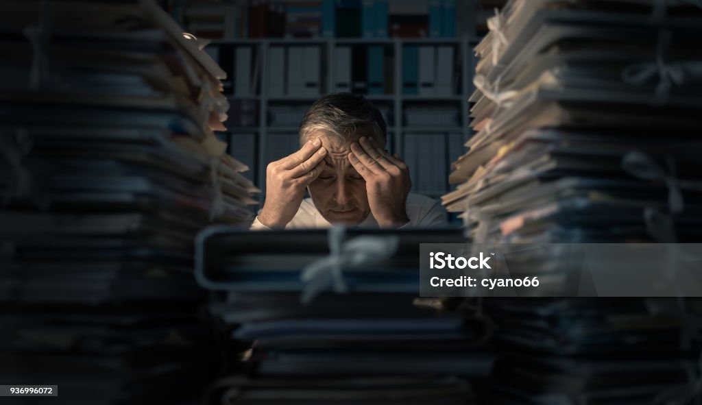 Desperate businessman working late Desperate businessman working in the office late at night and overloaded with work, his desktop is covered with paperwork: business management and deadlines concept Working Late Stock Photo