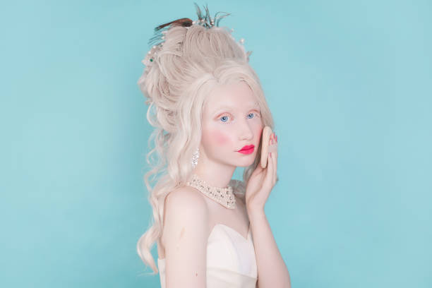 a blond woman with a beautiful luxurious rococo haircut in a white dress on a blue background. girl with puff for powder in hand - powder puff imagens e fotografias de stock