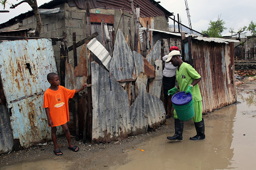 Cap Haitien, Haiti - March 16, 2018: A young boy watches a SOIL sanitation worker collect a full bucket from a client with an EkoLakay Ecosan home composting toilet in the Aviasyon neighborhood, which is subject to frequent flooding. SOIL provides and services the toilets, which provide the basic elements, human waste and shredded vegetable matter, of compost that will, once completed and tested for pathogens, be used to grow fruits and vegetables.