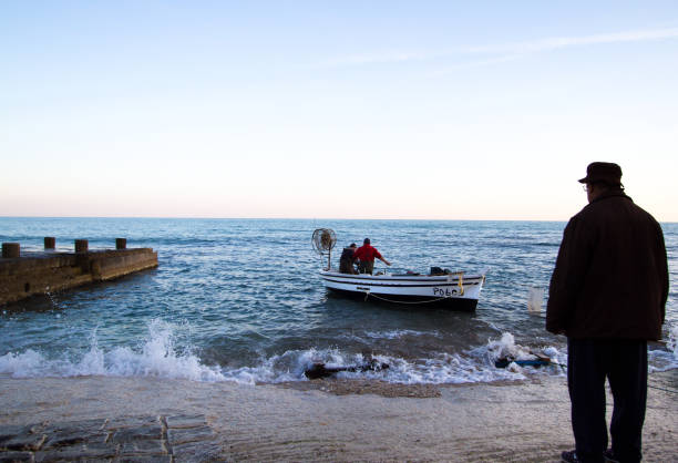 Sampieri (Ragusa), Sicily: Two fishermen in a boat at dusk approaching Sampieri, Sicily, a small fishing village in Ragusa Province, as a senior man on shore watches. Copy space in the sky.