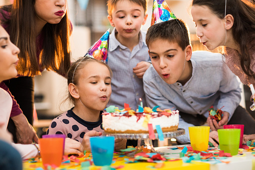 Group of children and an adult woman standing around the cake wearing party hats and feeling positive