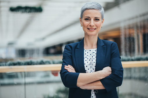 Portrait of successful female boss in business suit, mature African American woman smiling and looking at camera with crossed arms, businesswoman happy with achievement at workplace inside office.