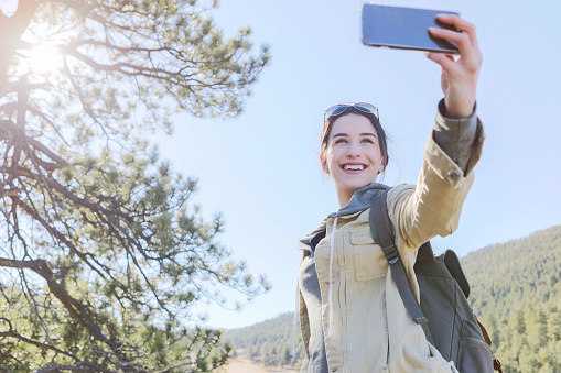 Attractive young Caucasian woman uses smart phone to take selfie while hiking in the mountains.