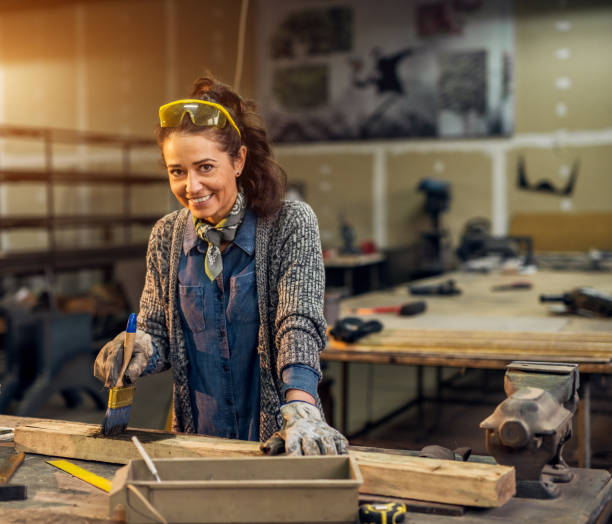 portrait view of happy attractive hardworking middle aged professional female worker painting wood with black colour in the sunny workshop or garage. - restoring art painting artist imagens e fotografias de stock