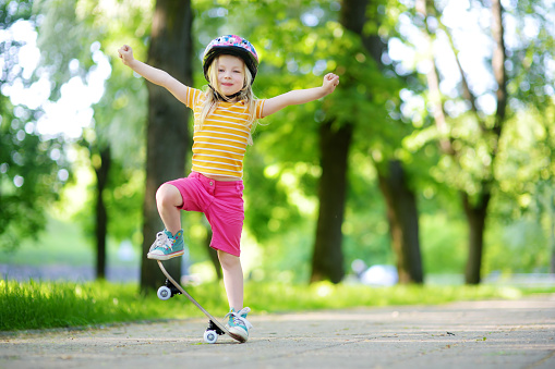 Pretty little girl learning to skateboard on beautiful summer day in a park. Child enjoying skateboarding ride outdoors.