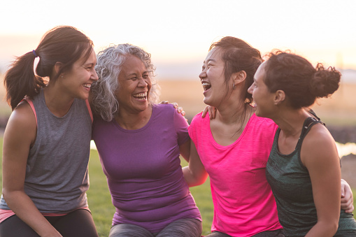 Four ethnic women of varying ages hug each other and laugh heartily after an intense workout together outside on a summer evening.