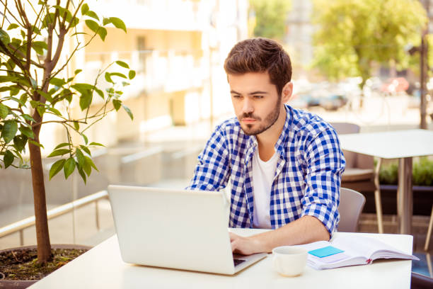 istock Young handsome bearded brunet is browsing on his laptop, sitting outdoors on a summer day, with note pad and cup of coffee, nice city view behind 937239658
