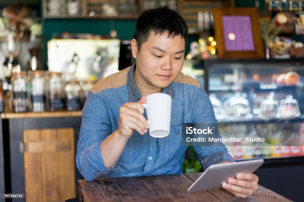 Content man reading article and drinking coffee Content Asian man reading article and drinking coffee in cozy cafe. Pleased handsome young man relaxing in public place. Coffee house concept Adult Stock Photo