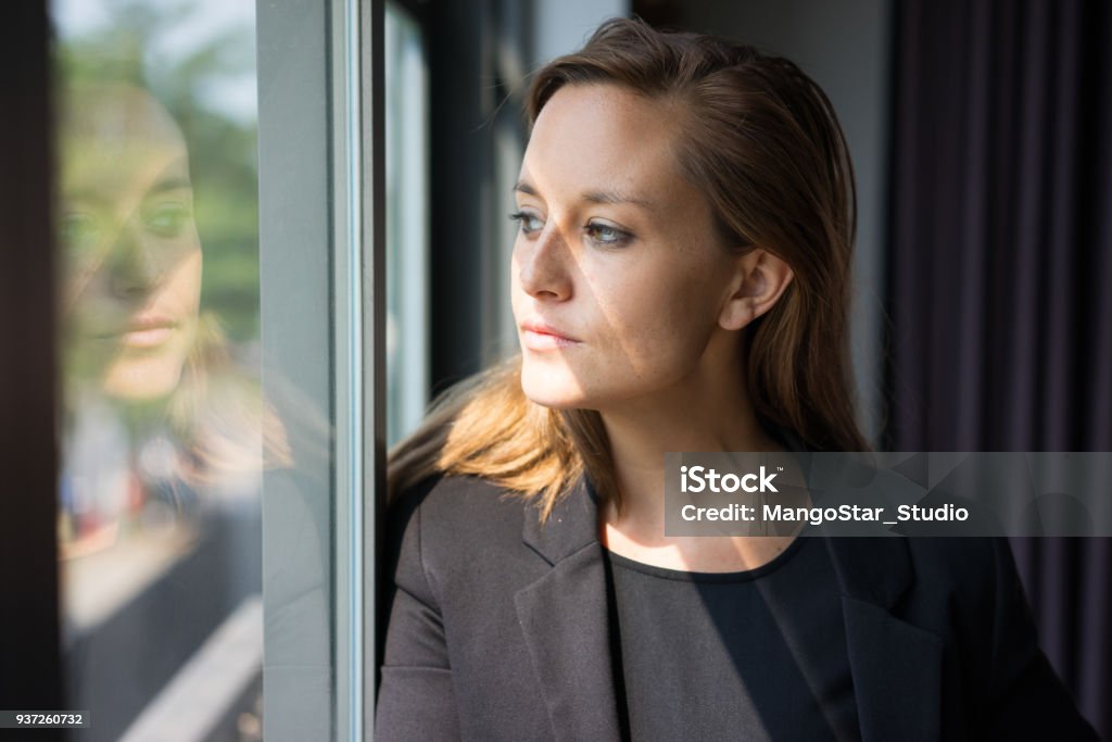 Pensive Business Lady Looking Through Window Closeup portrait of pensive young beautiful brown-haired woman looking through window. Contemplation concept. Front view. Women Stock Photo