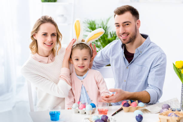 istock Celebrating family with daughter playing with Easter bunny ears by table with painted eggs 937265738