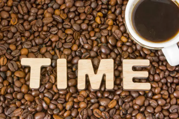 Word Time composed of large letters with wooden structure is located on the background of coffee beans with a cup of brewed coffee. Symbol large drinking coffee time
