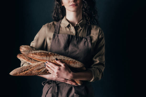 cropped shot of woman in apron holding french baguettes in hands isolated on black cropped shot of woman in apron holding french baguettes in hands isolated on black bread bakery baguette french culture stock pictures, royalty-free photos & images