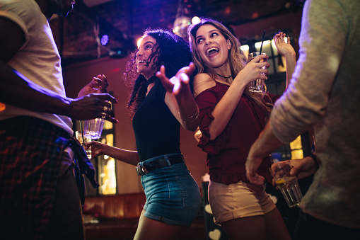 Happy young women dancing and partying with male friends at nightclub. Group of friends enjoying at bar with drinks.