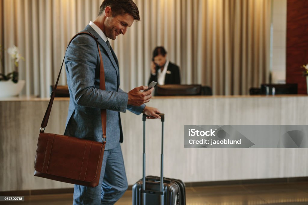 Businessman arriving at hotel lobby Businessman standing in hotel lobby with suitcase and using his mobile phone. Male business traveler in hotel hallway with smartphone and luggage. Business Travel Stock Photo