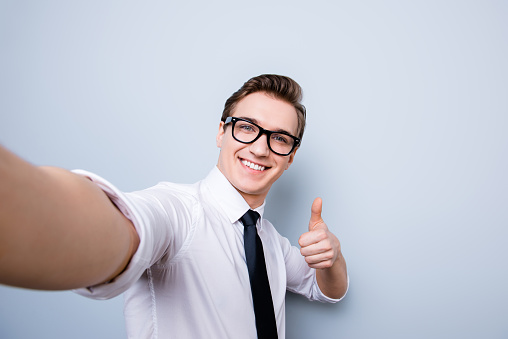 Successful excited young guy in glasses and formal wear is making selfie shot on camera, standing on a pure background, showing thumb up sign, smiling