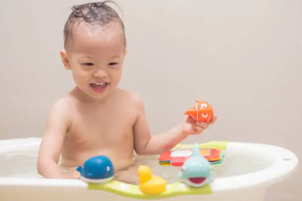 Photo of Asian 18 months / 1 year old toddler baby boy child taking a bath at home, Smiling kid having fun in bath time playing with colorful rubber toy,
