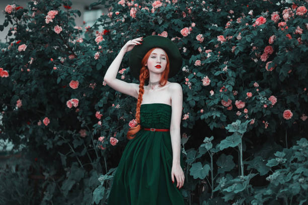 das rothaarige mädchen in einem langen kleid und vintage grünen hut posiert vor dem hintergrund der pfirsich rosen. romantische dame mit einem roten zopf und roten lippen im stil der aufklärung. blumengarten - braided women sensuality portrait stock-fotos und bilder