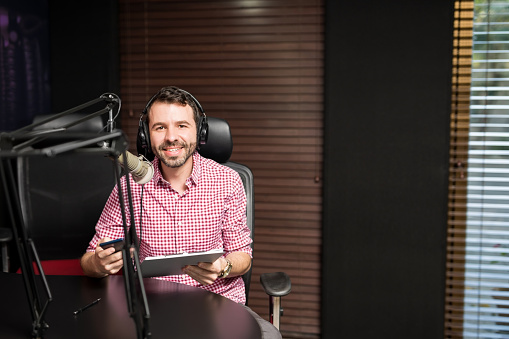 Portrait of smiling hispanic male radio presenter with clipboard hosting a show at radio station