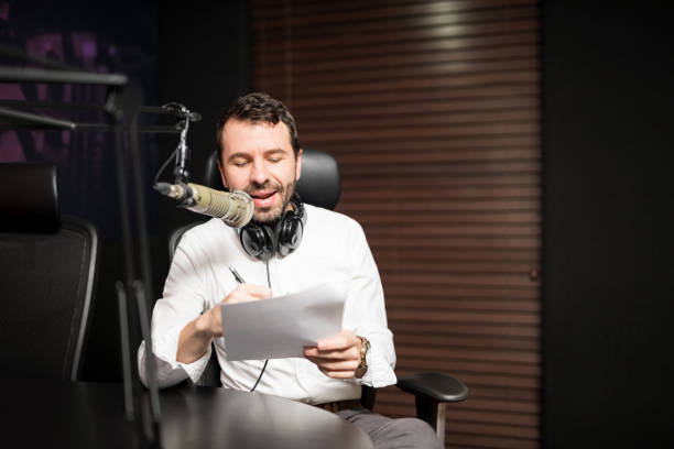 Handsome male radio host in studio Portrait of young male radio host going live on air talking in microphone holding a script paper in studio commentator photos stock pictures, royalty-free photos & images