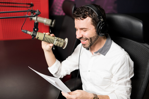 Smiling young male radio presenter with headphones reading a script and talking into a mic in a radio station