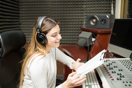 Attractive woman with clipboard in hand sitting in soundproof room at radio station and hosting a talk show live