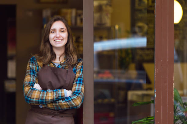 hermosa vendedora joven mirando a cámara y apoyado contra el marco de la puerta de una tienda de orgánico. - puerta negocio fotografías e imágenes de stock