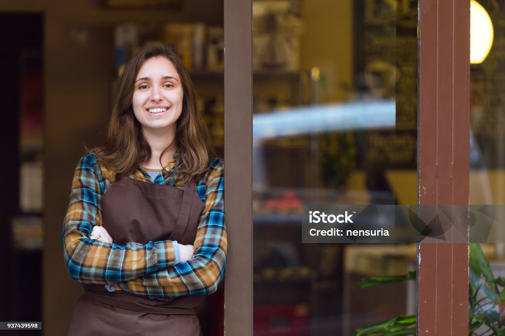 Beautiful young saleswoman looking at camera and leaning against the door frame of an organic store. Portrait of beautiful young saleswoman looking at camera and leaning against the door frame of an organic store. Store Stock Photo