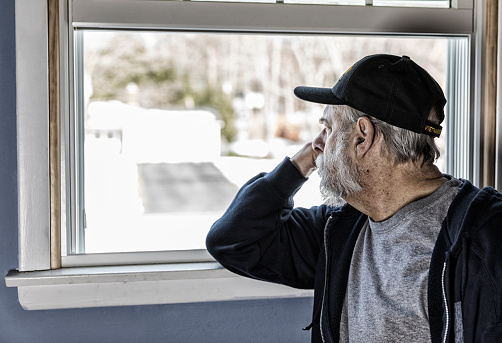 An authentic 67 year old United States Navy Vietnam War senior adult man military veteran is sitting at home looking away through an open window. The veteran is wearing an inexpensive, non-branded, generic, souvenir shop replica Vietnam veteran commemorative baseball hat style cap. Potential image uses to illustrate or highlight military veterans' problems and opportunities returning to civilian life; veterans' healthcare, medicine and mental illness concepts including PTSD (post traumatic stress disorder), sadness, anxiety, coping, emotional stress, nostalgia, melancholy, loneliness, therapy, recovery, assistance, renewal, raison d'etre, caring, etc.
