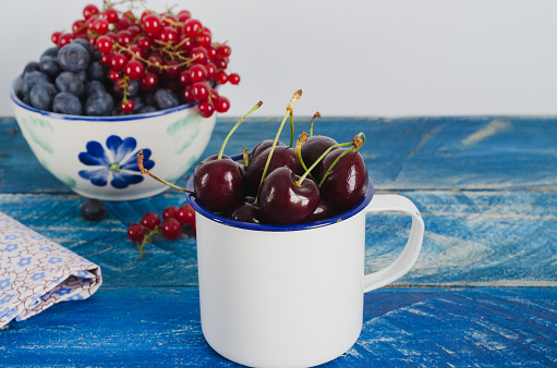 Red cherries and cranberries in white bowl on aged wooden background of blue color.