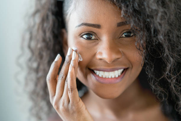 istock Beautiful black woman applying moisturizer on her face while looking at camera smiling 937487270