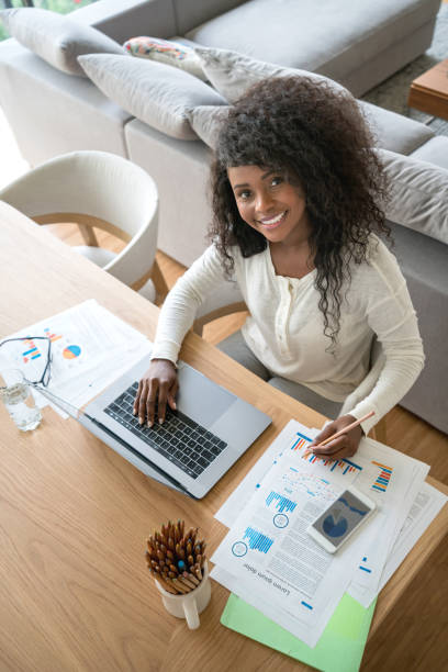 istock Pretty black young woman working from home using her laptop, smartphone and documents but looking at camera smiling 937487418