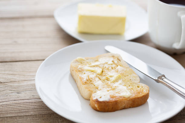 sclice of toasted bread with a spread of butter on a white plate, all on rustic wooden board, a piece of butter in the background