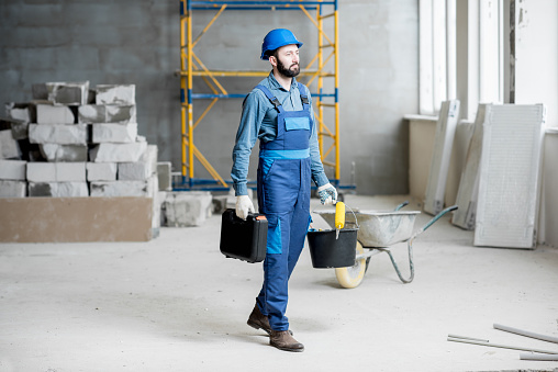 Builder in working uniform with protective helmet standing with instruments at the construction site indoors