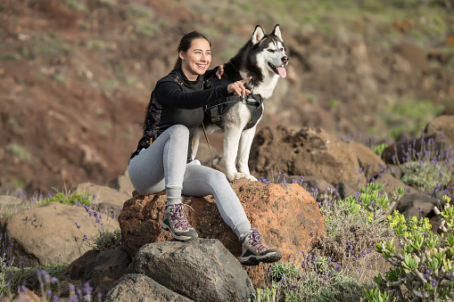 Cheerful woman in sportive outfit sitting with Husky on stone and pointing away in excitement.