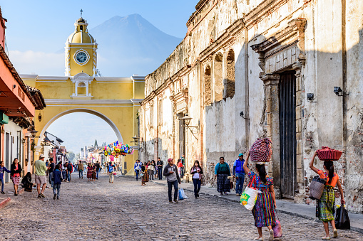 Locals & tourists walk in popular tourist street with Santa Catalina arch & ruins in colonial town & UNESCO World Heritage Site against backdrop of Agua volcano
