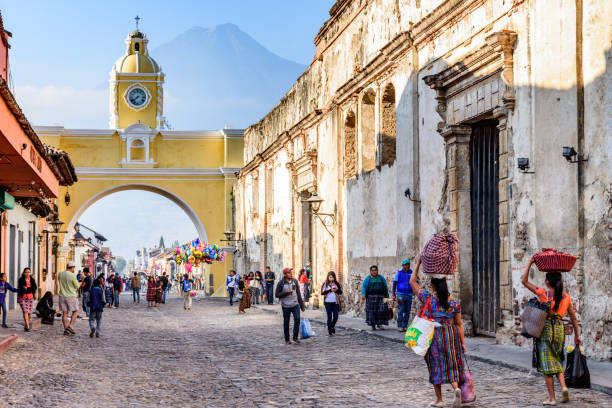 Street with Santa Catalina arch, ruins & volcano, Antigua, Guatemala Locals & tourists walk in popular tourist street with Santa Catalina arch & ruins in colonial town & UNESCO World Heritage Site against backdrop of Agua volcano agua volcano photos stock pictures, royalty-free photos & images
