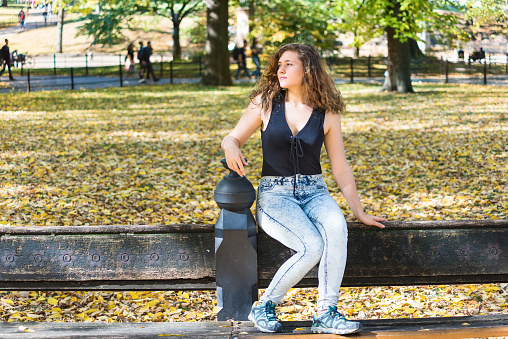 Manhattan New York City NYC Central park with trees, young hipster millennial woman sitting on bench, many fallen leaves in autumn fall season with yellow vibrant saturated foliage