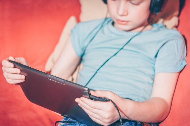 istock Red-haired boy sitting on sofa playing games on tablet 937704572