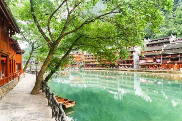 Scenic embankment of the Tuojiang River (Tuo Jiang River) in Phoenix Ancient Town (Fenghuang County), China. Traditional Chinese wooden riverside buildings among green trees.