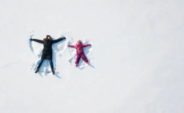 Photo of Child girl and mother playing and making a snow angel in the snow. Top flat overhead view