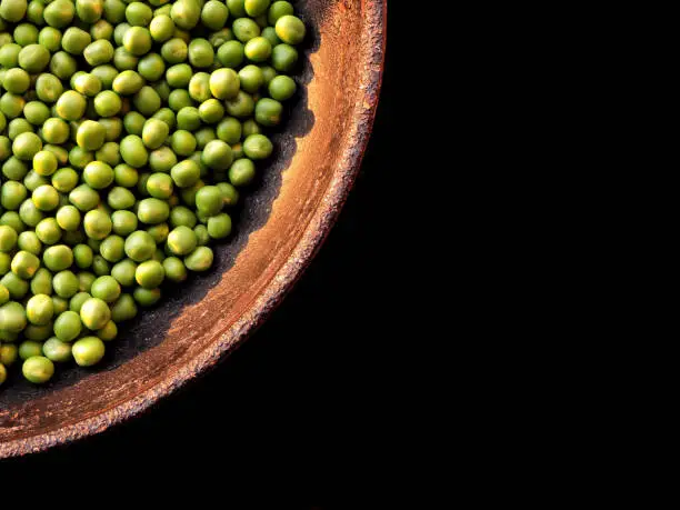 collection of shelled fresh green peas shot from top with evening sunlight reflecting on it while being placed in a brown bowl at top left corner of the image on a black background